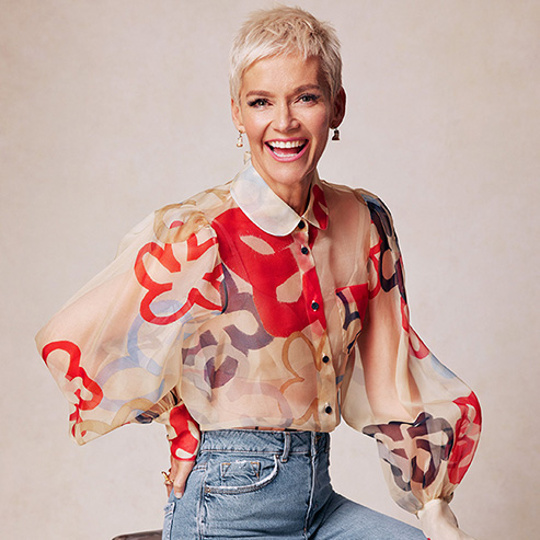 Jessica Rowe wearing a colorful shirt and jeans, sitting on a chair smiling