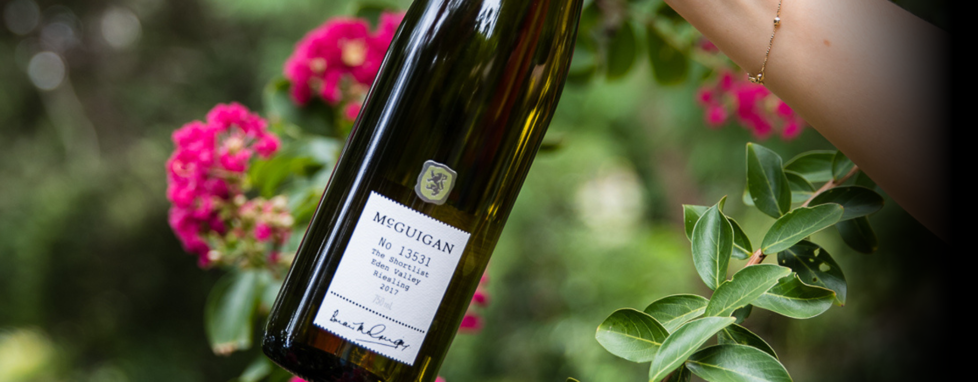 McGuigan Riesling bottle in the air in front of green bushes and pink flowers