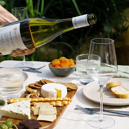 Bottle of McGuigan Mastercraft Chardonnay being poured into a glass with a charcuterie board containing crackers and cheese