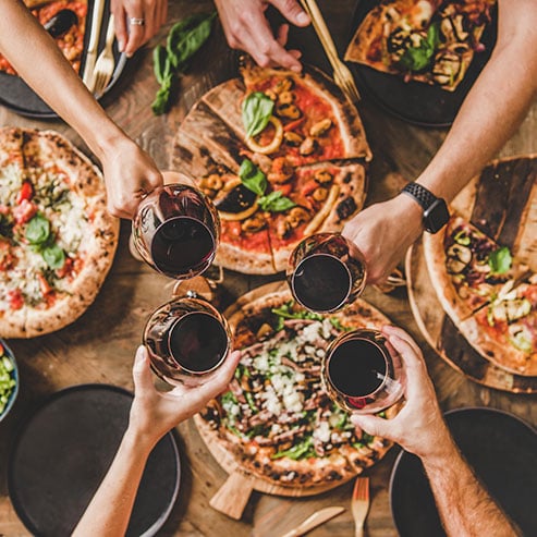 Four individuals holding wine glasses with assorted pizzas in the background